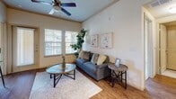 This single-floor residence offers a thoughtfully designed layout encompassing a living room, dining area, kitchen, two bedrooms, and two bathrooms. The living room features wood-look flooring, crown molding, a ceiling fan, and large windows fitted with blinds that allow ample natural light while maintaining privacy. The kitchen is equipped with warm wood cabinetry, dark countertops, black appliances including a range, microwave, and refrigerator, along with a peninsula breakfast bar illuminated by pendant and track lighting. Adjacent to the kitchen, the dining nook includes a built-in desk area with open shelving above, providing a functional workspace alongside the dining space. The primary bedroom is carpeted and includes a ceiling fan and a window with exterior views, while a walk-in closet and an en-suite bathroom with a glass-enclosed shower, dark-topped vanity, and wood cabinetry complete the suite. The second bedroom is also carpeted with natural light from a window. The second bathroom features a soaking tub with tile surround, matching dark-top vanity, and tile flooring. Throughout, the warm neutral palette creates a cohesive, comfortable ambiance.