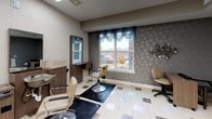 This professionally designed salon space offers a polished and comfortable environment for personal care services. The space is finished with a sophisticated aesthetic, combining two distinct wall treatments — a soft, textured linen-toned wallcovering on one side and a bold, embossed floral-patterned gray wallcovering on the other — creating a dynamic yet cohesive interior. The flooring is a standout feature, consisting of light-toned planks accented with inlaid bronze and silver square tiles arranged in a decorative pattern that adds elegance throughout.

The active styling area is equipped with professional hydraulic salon chairs, a dedicated shampoo station with a built-in basin and wood-toned cabinetry, and a vanity mirror. A large window dressed with layered teal valance curtains floods this area with natural light and offers views of the surrounding exterior. Adjacent to the styling zone is a comfortable waiting area furnished with upholstered lounge chairs and a sculptural side table, lit by two striking gold drum pendant lights. Wall art and decorative metal accents contribute to the refined ambiance. Large recessed panel ceiling lights provide bright, even illumination throughout. The space is designed to feel both functional and inviting, balancing professional utility with a warm, stylish atmosphere.