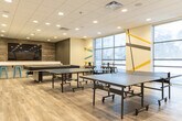 Located at 1700 Simcoe St N in Oshawa, Ontario, this energetic recreational amenity space is designed to foster social interaction and active play. The open-concept layout is finished with light wood-look flooring throughout, complemented by a clean drop-ceiling with recessed lighting that evenly illuminates the entire area.

A defining design feature is the bold, large-scale geometric mural that spans multiple walls, incorporating striking tones of yellow, teal, grey, and black to create a dynamic and visually stimulating environment. Large windows along the exterior wall flood the space with natural light and offer views of the surrounding neighbourhood.

The games area is well-equipped with multiple full-size ping pong tables and a shuffleboard or billiards table, providing ample room for competitive play. A long, white bar-height counter lined with bright teal metal stools offers spectator seating and a casual gathering spot. A wall-mounted flat-screen TV is set against a wood-panelled accent wall, adding a media viewing element to the room.

A comfortable lounge zone is anchored by a generously sized teal sectional sofa arranged around a round wooden coffee table, creating an inviting area for relaxation. High-top tables and stools near the windows provide additional seating. The space combines a modern, youthful aesthetic with practical, community-focused amenities.