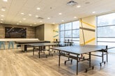 This 1,090 square foot space is a contemporary recreational area designed for versatility and engagement. The room showcases a modern aesthetic with a striking balance of functionality and style. Large windows flood the area with natural light, offering views of the outdoors and creating an open, airy atmosphere.

The space is characterized by its unique wall designs, featuring bold geometric patterns in yellow, blue, and gray, which add visual interest and energy to the white walls. The floor is covered with high-quality wood-look vinyl planks, providing durability and warmth.

A standout feature is the large, L-shaped teal sectional sofa, offering ample seating for relaxation and socialization. Complementing this is a round wooden coffee table, perfect for casual gatherings or individual use.

The room is equipped for various activities, including table tennis tables for recreational sports. A bar-height counter with sleek blue stools provides additional seating and a space for casual dining or work. 

The versatile layout includes open floor areas that can accommodate exercise mats or other temporary setups, enhancing the room's multi-functionality. Recessed lighting throughout ensures even illumination, while a mounted TV screen on a feature wall adds entertainment options.

Overall, this space seamlessly blends comfort, style, and practicality, creating an inviting environment for diverse recreational and social activities.