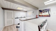 Located at 126 W Ahwanee Ave in Sunnyvale, California, this shared laundry facility is a well-organized, single-room space designed for efficient, everyday use. The room features light wood-look flooring and crisp white walls, giving it a clean and functional aesthetic throughout.

Multiple front-loading washing machines and stacked dryers are arranged along the walls, providing ample laundry capacity for residents. Dark granite-topped folding counters run along the perimeter, offering convenient surfaces for sorting and folding laundry. The counters are built at a comfortable standing height, maximizing usability within the compact footprint.

The ceiling is finished with white wood planking and exposed structural beams, adding a subtle rustic character to the otherwise modern interior. Flush-mounted and skylight-style light panels illuminate the space evenly, supplemented by natural light from exterior-facing doors on opposite ends of the room that allow for ventilation and easy access.

A fire extinguisher is mounted on the wall, and a security camera is positioned in the corner, ensuring safety throughout the space. Decorative touches, including a tropical beach artwork, add a welcoming feel. Informational signage is posted to guide users. The space is well-maintained and thoughtfully laid out to serve its practical purpose with ease and comfort.