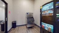 This single-floor utility room serves as a convenient amenity space, designed to provide easy access to refreshments and ice. The room is outfitted with a stainless steel commercial-grade ice machine equipped with a large storage bin, capable of meeting high-volume demand. Alongside it stands a multi-tier vending unit offering a selection of snacks and packaged goods, as well as a separate beverage vending machine stocked with canned drinks.

The space features wood-look luxury vinyl plank flooring throughout, complemented by neutral gray walls and dark blue baseboard trim that lend a clean, modern aesthetic. A branded entry mat bearing the property name is positioned near the doorway, reinforcing the building's identity. A small waste bin is conveniently placed within the room for easy disposal.

A glass-paneled door provides entry and visibility into an adjacent area, suggesting the room is part of a larger amenity corridor. The overall layout is functional and efficiently organized, with equipment positioned to allow comfortable movement within the space. Subtle recessed lighting keeps the room well-illuminated, contributing to a tidy and practical environment suited for regular, high-traffic use by building occupants.