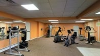 This fitness room offers a well-equipped workout space designed to support a variety of exercise routines. The room features warm orange walls that create an energizing atmosphere, complemented by a drop ceiling with recessed panel lighting that ensures consistent, even illumination throughout the space. The floor is covered in durable, low-pile carpeting suited for gym use.

The equipment selection includes multiple cardio machines — notably an elliptical trainer and a recumbent stationary bike — as well as a freestanding heavy bag for boxing or martial arts training. A multi-station cable and weight machine provides options for a range of strength training exercises. The layout is thoughtfully arranged to allow comfortable movement between stations.

A wall-mounted mirror near the entrance enhances the sense of space and allows users to monitor their form during workouts. A whiteboard is also visible on one wall, which may serve for workout tracking or instructional purposes. White-trimmed doorways provide access to adjacent areas. Overall, the space is functional, practical, and well-suited for regular fitness use.