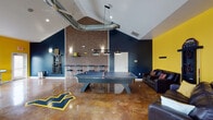 This single-floor community amenity building is thoughtfully divided into distinct functional areas, each with its own character and purpose. The front entry features a reception desk set on terracotta tile flooring beneath vaulted ceilings painted in cool gray-blue tones, offering a welcoming first impression with access to adjoining rooms.

The standout feature of the space is a generously sized game room with soaring vaulted ceilings and exposed industrial ductwork that adds an urban edge. Polished stained concrete floors in rich amber tones anchor the room, which is painted in bold navy blue and vivid yellow — an energetic color palette reinforced by a large logo inlaid directly into the floor. The game room is equipped with a ping pong table and a pool table, both suspended beneath modern pendant lighting. A brick accent wall, wall-mounted cue rack, bar-height seating, and leather lounge sofas complete the social atmosphere.

Adjacent to the game room is a seating lounge with leather armchairs and tile flooring, offering a quieter retreat. The business center features dramatic floor-to-ceiling angled windows that flood the carpeted room with natural light, and is outfitted with multiple desktop computer workstations for resident use.