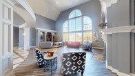 This expansive single-floor space offers an impressive blend of classical architectural detailing and comfortable, functional design across nearly 3,000 square feet. The centerpiece of the main living area is a dramatic vaulted ceiling that soars overhead, paired with a towering wall of arched, multi-pane windows that floods the room with abundant natural light and frames views of the surrounding landscape. Rich hardwood-style flooring runs throughout the living area, anchored by a stately white fireplace with an ornate mantel and marble surround — a true focal point of the room. Decorative white columns with detailed millwork define transitional spaces, while wall sconces and a gold chandelier add warmth and elegance. The lounge area features built-in media niches flanked by decorative alcoves, providing both function and visual interest. A reception desk area with wood flooring offers a professional touch, while an inlaid tile foyer with marble accents provides a grand entry. A separate kitchen and dining area includes wood cabinetry, bar-height seating, a ceiling fan, and tile flooring, making it well-suited for gatherings and events. Throughout, a consistent cool blue-gray color palette ties the spaces together, creating a refined and welcoming atmosphere.