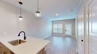 This single-floor unit showcases a thoughtfully designed open-concept layout with a clean, contemporary aesthetic. Light-toned luxury vinyl plank flooring flows throughout the main living areas, complementing the neutral wall palette and crisp white trim.

The kitchen is a standout feature, equipped with medium-toned wood cabinetry accented by matte black hardware, stainless steel appliances including a range, microwave, and refrigerator, and bright white quartz countertops. A subway tile backsplash adds a refined touch, while a kitchen island with pendant globe lighting serves as both a functional workspace and a visual centerpiece that connects seamlessly to the living area.

The living space is bright and open, benefiting from a large window and glass-paneled exterior door fitted with blinds, allowing for ample natural light and outdoor views. The bedroom features plush carpeting, a ceiling fan, and a window with blinds providing privacy and light control.

The bathroom offers a dual-sink vanity with white quartz countertops, wood-tone cabinetry, matte black fixtures, and a large dark-framed mirror with overhead vanity lighting, creating an elegant and functional space. Recessed lighting and ample closet access complete this well-appointed unit.