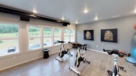 This single-level fitness center is thoughtfully designed to support a wide range of workout routines in a clean, contemporary setting. The main exercise area is anchored by a row of Spinning-brand stationary bikes arranged to face a large projection screen, making it ideal for guided cycling classes or on-demand workout sessions. Exposed black ductwork runs along the ceiling, lending the space a modern industrial aesthetic that contrasts with the light wood-look flooring and crisp white walls throughout.

Natural light fills the room through a generous bank of large windows that offer views of lush greenery outside, creating an open and energizing atmosphere. A full-length mirror along one wall enhances the sense of space and supports stretching or bodyweight training. Comfortable seating, including mustard-yellow ottoman-style seats and sleek black side tables, provides a place to rest between sessions.

A dedicated equipment zone houses a well-stocked rack featuring stability balls, medicine balls, a BOSU balance trainer, and a set of dumbbells, catering to functional and strength training needs. A wall-mounted flat-screen display adds a multimedia element to this area. The facility also includes an adjacent room visible through a glass-paneled door, recessed lighting throughout, and waste disposal stations, ensuring a practical and well-appointed workout environment.