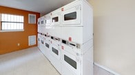 This shared laundry facility is a well-equipped, single-floor space designed to serve a communal setting with efficiency and practicality. The main laundry area is immediately recognizable by its bold orange accent wall, which adds a vibrant visual contrast to the rows of white top-loading washing machines and stacked dryers lining the walls. The machines are clearly numbered for easy identification, and a payment system is integrated for seamless operation. A large white folding table with hairpin legs provides a dedicated surface for sorting and folding laundry. The space is brightened by multiple barred windows that allow natural light to filter in, keeping the environment feeling open and airy. The light-toned vinyl tile flooring is easy to maintain and contributes to the clean, utilitarian aesthetic throughout. An adjoining waiting area features neutral beige walls and a bench with molded white seats, offering a comfortable spot to wait. A window in this section looks out toward greenery, adding a touch of calm to the functional space. A small nook with additional seating and a waste bin provides further convenience. Overhead fluorescent lighting ensures the entire facility remains well-lit at all times, making this laundry room a reliable and functional amenity.