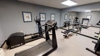This dedicated fitness room offers a comprehensive workout environment designed to accommodate a range of exercise needs. The space is thoughtfully laid out to maximize functionality, featuring a combination of cardio and strength training equipment. Cardio machines include a treadmill and an elliptical trainer, both positioned for easy access and use. A large wall mirror spans one side of the room, visually expanding the space and allowing users to monitor their form during exercise. On the strength training side, a weight bench and a rack with free weights provide options for resistance workouts. The light-colored carpet flooring and soft grey walls create a clean, neutral backdrop, while bright overhead lighting ensures the room is well-lit at all times. Instructional exercise posters are mounted along the walls, offering guidance on proper equipment usage for cycling, elliptical, and treadmill workouts. The overall ambiance is practical and focused, providing an organized, distraction-free setting suited for fitness-oriented use. The single-floor layout ensures easy accessibility throughout the space.