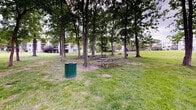 Located in Laurel, Maryland, this outdoor picnic area offers a welcoming communal green space designed for leisure and outdoor dining. The grounds are characterized by a generous expanse of well-maintained lawn, dotted with tall, mature trees that provide ample natural shade throughout the area. Beneath the tree canopy, several wooden picnic tables are arranged to accommodate group gatherings, complemented by charcoal grills conveniently positioned nearby for outdoor cooking. Waste receptacles, including a distinctive green trash can, are thoughtfully placed throughout the space to support cleanliness and ease of use. The terrain features a gentle grassy slope, lending a relaxed, park-like atmosphere to the setting. The open layout allows for flexible use of the grounds beyond the designated picnic zones. The surrounding backdrop includes a multi-story residential building with balconies visible through the tree line, situating the picnic area within a larger residential community. The combination of mature tree coverage, open green space, and practical amenities such as seating and grills makes this an inviting and functional outdoor area suited for casual gatherings, community events, and everyday outdoor relaxation.