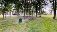 Located in Laurel, Maryland, this outdoor picnic area offers a welcoming communal green space designed for leisure and outdoor dining. The grounds are characterized by a generous expanse of well-maintained lawn, dotted with tall, mature trees that provide ample natural shade throughout the area. Beneath the tree canopy, several wooden picnic tables are arranged to accommodate group gatherings, complemented by charcoal grills conveniently positioned nearby for outdoor cooking. Waste receptacles, including a distinctive green trash can, are thoughtfully placed throughout the space to support cleanliness and ease of use. The terrain features a gentle grassy slope, lending a relaxed, park-like atmosphere to the setting. The open layout allows for flexible use of the grounds beyond the designated picnic zones. The surrounding backdrop includes a multi-story residential building with balconies visible through the tree line, situating the picnic area within a larger residential community. The combination of mature tree coverage, open green space, and practical amenities such as seating and grills makes this an inviting and functional outdoor area suited for casual gatherings, community events, and everyday outdoor relaxation.