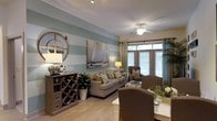 Located at 2945 Midtown Way in Wilmington, North Carolina, this single-floor residence offers a thoughtfully designed layout with a welcoming coastal aesthetic. The living area serves as the centerpiece of the space, featuring soft sage-and-cream horizontal striped accent walls that evoke a relaxed, seaside atmosphere. A ceiling fan overhead provides both function and charm, while large windows allow generous natural light to fill the room, framing views of the outdoors with elegant draping curtains. The living area is anchored by a comfortable neutral-toned sofa accented with patterned throw pillows, complemented by a cozy armchair that creates an inviting seating arrangement. A rustic wooden wine rack credenza adds warmth and character, doubling as a stylish storage and display piece. Nautical-themed artwork adorns the walls, reinforcing the coastal design narrative throughout. The dining area flows seamlessly from the living space, featuring upholstered seating and a well-appointed table ideal for gatherings. Light-toned flooring runs throughout, unifying the open-concept layout and enhancing the bright, airy feel of the home. Overall, this residence combines practical design with a refined coastal charm that is both comfortable and visually appealing.