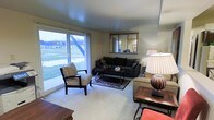 This single-floor space presents a functional and comfortably arranged living area that balances natural light with a warm, inviting atmosphere. The living room serves as the centerpiece of the space, featuring wall-to-wall carpeting in a neutral tone that contributes to its cozy character. A standout element is the large sliding glass window along one wall, which floods the room with natural light and frames views of the surrounding outdoor greenery and neighboring structures.

The living room is thoughtfully appointed with a dark upholstered sofa accented with decorative pillows, a striped armchair with a classic silhouette, and a wrought-iron coffee table resting atop a patterned area rug. An ornate framed mirror mounted on the wall adds depth and a touch of elegance to the room. Warm ambient lighting is provided by a generously sized table lamp with a pleated shade, complemented by additional side tables that offer practical surface space.

The overall ambiance is relaxed and traditionally styled, with furnishings that reflect a timeless aesthetic. The neutral wall tones and soft carpeting create a serene backdrop throughout. The single-floor layout ensures ease of movement and accessibility, making the space practical while retaining a lived-in, welcoming character throughout.