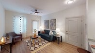 Located along West Tyvola Road in Charlotte, NC, this single-story residential unit offers a well-organized layout that balances functionality and comfort. The floor plan, visible through a 3D overview, reveals a logical flow between the living area, bedroom, kitchen, and bathroom, making efficient use of the available space.

The living room features light hardwood-style flooring, neutral white walls, and a ceiling fan overhead. A glass-paneled door allows natural light to filter in, creating a bright and welcoming atmosphere. The space accommodates a full seating arrangement comfortably.

The kitchen is outfitted with light wood cabinetry extending to the ceiling for ample storage, granite-style countertops, and a full suite of stainless steel appliances including a refrigerator, range, and over-the-range microwave. The wood-tone flooring continues throughout, maintaining visual consistency.

The bedroom is carpeted and features a ceiling fan with light fixture, a window with sheer curtains that invite natural light and outdoor views, and sufficient space for bedroom furnishings including nightstands and a dresser. Panel-style doors add a clean, classic detail throughout the unit.

The bathroom includes a granite-topped vanity with light wood cabinetry, a large mirror, and a full shower-tub combination, finished with hardwood-style flooring for a cohesive look.