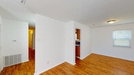 This single-floor unit offers a straightforward, functional layout with a clean, neutral aesthetic throughout. The main living and dining areas are finished with warm-toned hardwood-style flooring and crisp white walls, creating a bright and open atmosphere. Natural light filters in through windows, enhancing the airy feel of the space. The kitchen is accessible via a pass-through opening from the main living area and is equipped with warm wood cabinetry, dark granite countertops with a subtle tile backsplash, a double stainless steel sink, and black appliances including a refrigerator, electric range, and microwave. Ample upper and lower cabinet storage provides practical functionality. The bedroom features plush carpeting, a ceiling fan with light fixture, a window for natural light, and a closet with a built-in rod for storage. The full bathroom includes wood-toned cabinetry with a granite-topped vanity, a toilet, and a full tub and shower combination with a curved curtain rod. Hardwood-style flooring continues into the bathroom, tying together the overall design. A hallway connects the living areas to the private rooms, completing this compact and efficient layout.