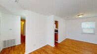 This single-floor unit offers a straightforward, functional layout with a clean, neutral aesthetic throughout. The main living and dining areas are finished with warm-toned hardwood-style flooring and crisp white walls, creating a bright and open atmosphere. Natural light filters in through windows, enhancing the airy feel of the space. The kitchen is accessible via a pass-through opening from the main living area and is equipped with warm wood cabinetry, dark granite countertops with a subtle tile backsplash, a double stainless steel sink, and black appliances including a refrigerator, electric range, and microwave. Ample upper and lower cabinet storage provides practical functionality. The bedroom features plush carpeting, a ceiling fan with light fixture, a window for natural light, and a closet with a built-in rod for storage. The full bathroom includes wood-toned cabinetry with a granite-topped vanity, a toilet, and a full tub and shower combination with a curved curtain rod. Hardwood-style flooring continues into the bathroom, tying together the overall design. A hallway connects the living areas to the private rooms, completing this compact and efficient layout.