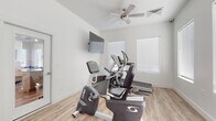 This dedicated fitness center is designed to support a comprehensive workout experience within a single, well-organized space. The room features light wood-style flooring throughout, crisp white walls, and ample natural light streaming in through multiple windows fitted with horizontal blinds, creating a clean and energizing atmosphere. A ceiling fan overhead helps maintain comfortable airflow during workouts.

The cardio zone is equipped with a Cybex recumbent exercise bike, a Sole elliptical trainer, and a treadmill, all arranged to allow easy access and movement between machines. A wall-mounted flat-screen television is positioned for viewing during cardio sessions. For strength training, the space includes a Hoist multi-functional cable machine with dual weight stacks, offering a wide range of resistance exercises. A dedicated free weights area houses a full dumbbell rack with multiple weight increments alongside an adjustable workout bench, providing versatility for users of all fitness levels.

A glass-panel door on one side of the room allows direct access to the exterior, further enhancing ventilation and adding to the open feel of the space. The overall layout is thoughtfully arranged to maximize usability while keeping the environment uncluttered, making this fitness center a practical and inviting facility for daily exercise.