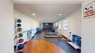 Located at 2112-A Criston Dr in Newport News, VA, this fitness facility offers a comprehensive workout environment designed to support a wide range of exercise routines. The space is laid out on a single floor and features a distinctive flooring combination of warm hardwood and durable rubber matting, providing designated zones for cardio and functional training activities.

The main exercise room is outfitted with a lineup of cardio equipment, including multiple treadmills, an elliptical trainer, a stationary bike, and a rowing machine. A dedicated strength training area features a cable machine, adjustable weight bench, and a rack of dumbbells, while a medicine ball storage rack adds versatility for functional workouts. Recessed ceiling lighting illuminates the space evenly, and multiple windows along the walls allow natural light to filter in, creating a bright and energizing atmosphere.

A wall-mounted mini-split HVAC unit ensures climate control throughout the room, while a mounted TV screen provides entertainment or instructional content. The entrance area is clean and functional, equipped with a foam roller and accessory storage rack, a waste bin, and a sanitation station, ensuring a hygienic environment for all users. The glass-panel entry door allows additional natural light into the space. Overall, the facility delivers a practical and well-organized setting for community fitness needs.