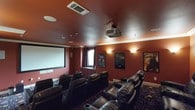 This dedicated home theater is designed to deliver a true cinematic experience from the comfort of home. The room is finished with rich, deep-red walls that set a dramatic and immersive atmosphere, complemented by crown molding and a matching ceiling that enhances the intimate, enclosed feel of the space. A ceiling-mounted projector points toward a large, framed projection screen anchored to the front wall, providing a commanding focal point for the room. Seating is arranged in tiered rows of plush, black leather power recliners, each equipped with built-in cup holders, ensuring optimal sightlines from every seat. Wall-mounted sconce lighting casts a warm, ambient glow along the side walls, while recessed ceiling lights and a dedicated HVAC vent ensure comfort during extended viewing sessions. The floor is covered in a richly patterned carpet that adds to the theatrical aesthetic. Framed movie posters adorn the walls, lending a classic cinematic character to the space. A small window with an arched detail provides a subtle architectural touch without compromising the room's light-controlled environment. This space is thoughtfully outfitted for an exceptional private screening experience.