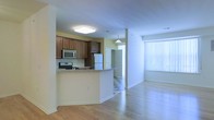 This single-floor space offers a clean, open-concept layout that connects the kitchen and living areas in a seamless, functional flow. The kitchen is outfitted with rich dark wood cabinetry, granite countertops, and stainless steel appliances including a gas range, built-in microwave, and refrigerator. A raised peninsula counter provides a natural divide between the kitchen and the adjoining living space, ideal for casual dining or meal prep. Light hardwood flooring runs throughout, adding warmth and continuity to the space. The living area is generously proportioned, bathed in natural light streaming through a large window dressed with vertical blinds. A pendant light fixture adds a subtle decorative touch near the entryway corridor, which features a classic checkered tile floor offering a distinct transition between spaces. Soft yellow-toned walls complement the light wood floors and white trim, creating a welcoming, airy ambiance throughout. The overall layout is practical and well-organized, making efficient use of every area while maintaining an open, comfortable feel.
