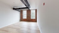 This thoughtfully renovated loft-style unit showcases a striking blend of historic industrial character and contemporary design. Exposed red brick walls serve as a defining architectural feature throughout, complemented by light hardwood flooring and crisp white walls that create a bright, airy atmosphere. Overhead, black industrial ductwork runs across the soaring ceilings, adding an authentic warehouse aesthetic that ties the space together.

The open living and bedroom areas benefit from generous natural light streaming through large, multi-pane windows that frame views of the surrounding neighborhood. Each room offers ample space and a clean, versatile canvas.

The modern kitchen is well-appointed with two-tone cabinetry — white uppers paired with warm wood-tone lowers — stone countertops, a subway tile backsplash, and a full suite of stainless steel appliances including a range, microwave, refrigerator, and dishwasher. A peninsula provides additional prep space and casual seating. A dedicated in-unit laundry area adds everyday convenience.

The unit includes two bathrooms, both finished with large-format neutral tiles. One features a glass-enclosed walk-in shower with a built-in seat and an adjacent vanity, while the second offers a similarly sleek, fully tiled wet room design. Together, these spaces deliver a polished, contemporary feel within a space rich with historic character.