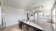 This well-appointed single-floor residence showcases a thoughtfully designed open-concept layout that seamlessly connects the kitchen and living areas. The kitchen is a standout feature, boasting granite countertops on an expansive island with a stainless steel sink, dark cabinetry with sleek hardware, tile backsplash, and a full suite of stainless steel appliances including a refrigerator, range, and microwave. Pendant lighting and a track lighting system add both function and style overhead, while light tile flooring flows throughout the main living spaces. The living area extends toward large windows dressed with horizontal blinds, allowing generous natural light to fill the room, and is equipped with a ceiling fan for comfort. A well-lit hallway connects the private quarters, featuring tile flooring and panel doors with brass hardware. The bedrooms are carpeted and bright, with large windows framing urban skyline views and ceiling fans overhead. The bathrooms are finished with granite-topped dark vanities, large mirrors, soaking tubs with tiled surrounds, and tile flooring. A spacious walk-in closet with dual hanging rods and shelving provides ample storage. The overall aesthetic is clean, modern, and cohesive throughout.