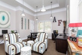 Located at 11170 Brunson Dr in Johns Creek, Georgia, this single-floor space functions as a well-appointed community clubhouse or leasing office designed to be both functional and inviting. The main lounge area is bathed in natural light streaming through a generous bank of tall windows fitted with white blinds, which extend nearly to the ceiling and offer views of surrounding greenery. Soaring ceiling heights amplify the sense of openness, while industrial-style pendant lighting adds a contemporary touch. The lounge is furnished with striped accent chairs and ottomans arranged around a central gathering area, complemented by a rustic, colorfully painted sideboard and a coffee station — making it an ideal space for welcoming visitors. Curated wall art and decorative twig arrangements contribute to the eclectic, warm ambiance throughout. A dedicated private office features a coffered ceiling, a large dark wood conference table, a full workstation with cabinetry, and ample natural light from sizable windows. An additional workstation area equipped with a computer and printer provides practical workspace. Warm neutral tones, patterned carpeting, and crown molding unify the interior design, while wide hallways connect the spaces seamlessly, creating a cohesive and professional environment.