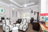 Located at 11170 Brunson Dr in Johns Creek, Georgia, this single-floor space functions as a well-appointed community clubhouse or leasing office designed to be both functional and inviting. The main lounge area is bathed in natural light streaming through a generous bank of tall windows fitted with white blinds, which extend nearly to the ceiling and offer views of surrounding greenery. Soaring ceiling heights amplify the sense of openness, while industrial-style pendant lighting adds a contemporary touch. The lounge is furnished with striped accent chairs and ottomans arranged around a central gathering area, complemented by a rustic, colorfully painted sideboard and a coffee station — making it an ideal space for welcoming visitors. Curated wall art and decorative twig arrangements contribute to the eclectic, warm ambiance throughout. A dedicated private office features a coffered ceiling, a large dark wood conference table, a full workstation with cabinetry, and ample natural light from sizable windows. An additional workstation area equipped with a computer and printer provides practical workspace. Warm neutral tones, patterned carpeting, and crown molding unify the interior design, while wide hallways connect the spaces seamlessly, creating a cohesive and professional environment.