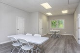 This single-floor space offers a clean, functional layout designed to accommodate a variety of professional uses. The interior is finished with light-toned luxury vinyl plank flooring throughout, lending a modern, cohesive appearance across the open rooms. Walls are painted in a warm neutral beige, creating a calm and professional atmosphere.

The drop-tile ceiling is equipped with recessed fluorescent panel lighting, ensuring consistent, even illumination throughout the space. A wall-mounted mini-split HVAC unit provides independent climate control, adding to the overall functionality of the environment.

Multiple white-framed windows allow natural light to filter into the rooms, offering views of surrounding greenery and outdoor surroundings. The windows contribute to an open, airy feel that complements the neutral interior palette.

The space includes multiple interior doors connecting separate areas, providing flexibility for private meetings or breakout sessions. The rooms are currently furnished with white folding tables and chairs, demonstrating their suitability as meeting or conference rooms. White panel doors with classic raised molding add a finished, polished detail throughout.

Overall, the space presents a tidy, well-maintained environment well-suited for professional, educational, or collaborative uses.