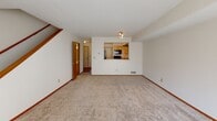 This two-floor residence offers a practical and well-organized layout designed for comfortable everyday living. The main level features a spacious living area with plush carpeting, neutral walls, and a sliding glass door fitted with vertical blinds that invites natural light and provides access to the outdoors. A pass-through window connects the living area to the kitchen, creating an open, communicative feel between the two spaces. The kitchen is equipped with warm honey-toned cabinetry, a stainless steel sink, laminate countertops, hardwood-style flooring, and full-size black appliances including a refrigerator, range, and dishwasher. A dedicated laundry room houses a full-size washer and dryer alongside a wire storage shelf overhead, adding significant convenience. The bathroom features a full soaking tub with white tile surround, a curved shower rod, a built-in linen tower with wood cabinetry, and a vanity with additional storage. Bedrooms are carpeted and bright, with large windows that allow natural light to filter in throughout the day. The primary bedroom includes dual closets offering ample storage. Consistent warm wood trim and neutral tones throughout create a cohesive and welcoming atmosphere across both floors.