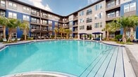 This striking residential community showcases a thoughtfully designed courtyard centered around a generously sized resort-style swimming pool with crystal-clear blue water. The multi-story building wraps around the outdoor space in a U-shaped configuration, creating a sense of enclosure and community while offering private balconies that overlook the amenity area below.

The exterior architecture features a bold combination of deep blue vertical cladding and gray siding, accented by large windows and black metal balcony railings that lend the complex a contemporary, upscale aesthetic. Tall palm trees are strategically placed throughout the courtyard, reinforcing a tropical, resort-like ambiance that complements the warm-climate setting.

The expansive pool deck is finished with large-format stone-look pavers and lined with modern sling-style lounge chairs paired with teal umbrellas, providing ample space for sunbathing and relaxation. A covered pergola structure on one side of the deck offers a shaded outdoor dining and gathering area, complete with seating and a built-in grill or fire feature — ideal for entertaining.

Tropical plantings, including ornamental grasses and colorful foliage, border the deck and soften the hardscape transitions. The overall design balances bold architectural character with inviting outdoor living, creating a vibrant communal space suited for an active, amenity-rich lifestyle.