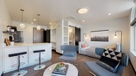 This single-floor studio unit showcases a thoughtfully designed open-concept layout that seamlessly integrates the living, sleeping, and kitchen areas into one cohesive space. Dark hardwood-style flooring runs throughout, grounding the bright white walls and creating a modern, urban aesthetic.

The kitchen is efficiently arranged in an L-shape, featuring white flat-front cabinetry, a white quartz countertop, an induction cooktop, stainless steel sink, dishwasher, and a wall-mounted utensil rail. Open dark shelving adds functional display space, while glass pendant lights above the breakfast bar lend a decorative touch. A wall-mounted mini-split HVAC unit ensures year-round climate control.

The living area benefits from large arched and rectangular windows that flood the space with natural light and offer views of the surrounding streetscape. A built-in open shelving unit provides additional storage and organization near the entryway.

A sliding black barn door leads to the bathroom, which features a subway-tiled walk-in shower, a pedestal sink, concrete-style flooring, and open built-in shelving for toiletries and linens. The sleeping area is integrated into the main living space, completing this compact yet well-appointed studio.