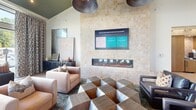 Located at 2020 Winters Eve Dr in Charlotte, NC, this thoughtfully designed community lounge offers an expansive, open-plan living area that masterfully blends contemporary elegance with inviting comfort. The single-floor space is anchored by a dramatic floor-to-ceiling tiled fireplace wall, complete with a wall-mounted flat-screen display above, serving as a striking focal point for one of several distinct seating arrangements.

Throughout the space, light wide-plank hardwood flooring contrasts beautifully with richly layered area rugs, while bold teal and olive-green ceiling treatments add architectural depth and visual intrigue. Oversized pendant lights in brushed metal descend dramatically from above, contributing to a sophisticated, curated ambiance.

Floor-to-ceiling windows along one full wall flood the interior with natural light and provide expansive views of the surrounding community. Patterned drapery panels frame the windows, adding warmth and texture. A built-in banquette with high-back seating and bistro-style tables offers an intimate dining nook near the windows.

Additional seating zones feature a mix of plush upholstered sofas, leather armchairs, and sculptural geometric coffee tables. A marble-effect accent wall with graceful arched sconce lighting anchors a relaxed lounge alcove. The overall ambiance is polished yet welcoming, reflecting a high standard of interior design throughout.