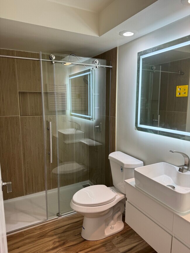 Baño moderno con ducha de cristal, tocador suspendido, lavabo sobre encimera y espejo con iluminación LED. - 328 N Allen St