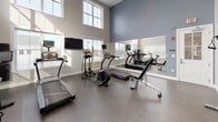 This fitness center offers a comprehensive workout environment designed to support a wide range of exercise routines. The space is characterized by soaring ceilings and expansive windows that line the upper walls, flooding the interior with natural light and creating an open, airy atmosphere. Durable speckled flooring throughout provides both safety and easy maintenance.

The cardio zone is well-appointed with treadmills, an elliptical trainer, a stationary bike, and a cycling bike, several of which are paired with mounted flat-screen televisions for entertainment during workouts. Large wall-mounted mirrors amplify the sense of space and allow users to monitor their form with ease.

The strength training area features a well-stocked dumbbell rack with a full range of free weights, an adjustable workout bench, and a multi-function cable machine, offering versatile options for resistance training. The layout is thoughtfully organized to ensure ample room to move between stations.

The entrance is marked by a branded welcome mat, and safety features including an emergency exit, fire alarm, and a mounted AED unit are clearly visible. Posted facility rules ensure a safe and organized environment for all users. Overall, the fitness center combines functionality with a clean, modern aesthetic.