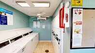 Located at 5601 FM-517 in Dickinson, Texas, this single-room laundry facility offers a practical and well-equipped space designed for efficient shared use. The room features light teal wood-paneled walls, neutral tile flooring, and a textured ceiling, giving the space a clean and functional aesthetic.

Multiple commercial-grade top-load washers and dryers are arranged along two walls, maximizing the use of the compact floor area and providing ample capacity for users. A built-in granite-top folding counter with a wall-mounted shelf above offers a convenient area for sorting and folding laundry, complemented by a trash receptacle below.

Climate comfort is addressed through both a ceiling fan and a wall-mounted mini-split air conditioning unit, ensuring a comfortable environment year-round. A window unit provides additional ventilation options. Natural light filters in through windows fitted with blinds, brightening the interior.

Safety is addressed with a clearly visible fire extinguisher mounted near the entrance. Informational signage and laundry tip posters are displayed on the walls, alongside lighthearted décor such as a "Have Loads of Fun" sign. A single exterior door with a window provides direct access to the outside. Overall, the facility is a tidy, purpose-built laundry space suited for community or shared residential use.