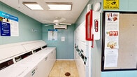 Located at 5601 FM-517 in Dickinson, Texas, this single-room laundry facility offers a practical and well-equipped space designed for efficient shared use. The room features light teal wood-paneled walls, neutral tile flooring, and a textured ceiling, giving the space a clean and functional aesthetic.

Multiple commercial-grade top-load washers and dryers are arranged along two walls, maximizing the use of the compact floor area and providing ample capacity for users. A built-in granite-top folding counter with a wall-mounted shelf above offers a convenient area for sorting and folding laundry, complemented by a trash receptacle below.

Climate comfort is addressed through both a ceiling fan and a wall-mounted mini-split air conditioning unit, ensuring a comfortable environment year-round. A window unit provides additional ventilation options. Natural light filters in through windows fitted with blinds, brightening the interior.

Safety is addressed with a clearly visible fire extinguisher mounted near the entrance. Informational signage and laundry tip posters are displayed on the walls, alongside lighthearted décor such as a "Have Loads of Fun" sign. A single exterior door with a window provides direct access to the outside. Overall, the facility is a tidy, purpose-built laundry space suited for community or shared residential use.