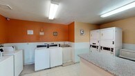This shared laundry facility is a single-floor space designed for practical, high-capacity use. The room is finished with a two-tone color scheme, featuring bold orange upper walls paired with lighter peach lower walls separated by a white wainscoting-style divider, creating a visually distinct and organized environment. Overhead fluorescent lighting ensures the space is well-lit throughout.

The facility is equipped with multiple top-load washing machines arranged along one wall, each fitted with coin-operated controls for convenient use. Opposite the washers, several stacked dryer units — numbered for easy identification — provide ample drying capacity. A utility sink is positioned between the machines, offering a practical handwashing or pre-treatment station.

A central granite-topped folding counter serves as the focal point of the room, providing generous surface area for sorting and folding laundry. The light tile flooring is easy to maintain and contributes to the clean, functional aesthetic of the space. Natural light enters through a window near the entry door, supplementing the overhead lighting.

Small framed artwork and decorative wall accents add a modest touch of character to the otherwise utilitarian space. The layout is thoughtfully arranged to allow comfortable movement between the machines and the central folding area.