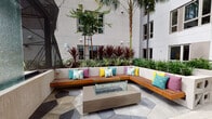 Located at 635 Robinson Ave in San Diego's Hillcrest neighborhood, this dynamic outdoor communal space is designed with a bold, modern aesthetic that encourages gathering and relaxation. The space is organized into distinct zones, each offering a unique social experience.

At the heart of one zone sits an L-shaped wood-slat bench with a sleek glass-enclosed fire pit table, surrounded by raised planters filled with lush ornamental grasses and dramatic red-leafed cordylines. Colorful throw pillows in teal, yellow, and purple add a lively, welcoming touch. Nearby, sculptural seating pieces in deep plum and vibrant red pod chairs with teal cushions create an eclectic conversation area.

A second zone opens onto a partially covered terrace showcasing a striking floor-to-ceiling textured glass water feature as its centerpiece. This area includes a fully equipped outdoor kitchen with a stainless steel grill, bar-height counter seating, and a dining area furnished with bold, multicolored cylindrical chairs in red, yellow, green, and blue. String lights and a geometric canopy overhead add ambiance, while sweeping views of neighboring palm trees and the San Diego skyline complete the setting. The patterned stone tile flooring ties the entire space together cohesively.