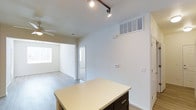 Located at 9444 East 108th Avenue in Henderson, Colorado, this single-floor residence offers a clean, functional layout designed for comfortable everyday living. The interior showcases a bright, open floor plan with light-toned luxury vinyl plank flooring that extends throughout the main living areas, contributing to a cohesive and airy feel.

The kitchen is equipped with a central island featuring a light countertop and dark cabinetry beneath, providing both additional workspace and storage. Overhead, a track lighting system delivers focused illumination, complementing the natural light that filters in from nearby windows. A ceiling-mounted HVAC vent ensures efficient climate control throughout the space.

The kitchen opens directly into the living area, which is outfitted with a ceiling fan and benefits from windows that invite natural light into the room. The seamless transition between the kitchen and living spaces makes the layout feel expansive and well-connected. A doorway visible from the main area leads further into the home, suggesting additional rooms beyond.

The walls throughout are finished in a neutral white palette, creating a versatile backdrop that suits a variety of interior styles. Overall, this home presents a well-maintained, move-in-ready environment with practical features and a straightforward, open design.