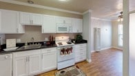 This single-floor residence offers a thoughtfully designed layout that balances functionality and comfort across its one bedroom and one bathroom. Crown molding details are present throughout, adding a refined architectural touch to the neutral, warm-toned walls.

The kitchen is well-equipped with white shaker-style cabinetry, dark granite countertops, and white appliances including an electric range with overhead microwave. A stainless steel sink anchors one end of the counter, while wood-look flooring ties the space seamlessly into the adjoining living area.

The living area is generously sized with plush carpeting and features a large picture window fitted with vertical blinds, allowing ample natural light to fill the room. A partial column with a mirrored accent panel serves as an architectural divider between the living and kitchen spaces, adding visual interest.

The bedroom is carpeted and well-lit by a pair of windows with blinds. It includes a built-in closet with sliding doors and a dedicated overhead storage niche, offering practical storage solutions.

The bathroom features tile flooring, a white vanity with dark countertop, a vessel-style sink, a large mirror, and a full tub-shower combination with a striped curtain.
