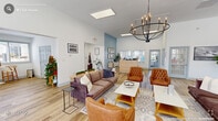 Located at Brookstone Lane Club in Fayetteville, NC, this single-floor community space offers a thoughtfully designed interior that balances comfort and functionality. The open layout is anchored by a spacious living room with soaring vaulted ceilings, light wood-look flooring, and abundant natural light streaming through a bank of large windows dressed with white blinds. A striking multi-arm candelabra chandelier serves as a focal point overhead, complementing the sophisticated seating arrangement of plush velvet sofas, tufted leather accent chairs, and contemporary coffee tables. Soft blue accent walls and curated wall art add warmth and visual interest throughout. Glass entry doors connect the living area to the exterior, enhancing the sense of openness. A dedicated lounge nook along the windowed wall provides additional seating with round bistro tables and wishbone chairs, bathed in natural sunlight. The kitchen features crisp white shaker cabinetry, granite countertops, stainless steel appliances including a full-size refrigerator, and a single-basin sink — all designed for convenient everyday use. A cozy dining area with a round white table and matching chairs sits adjacent, framed by a window with views of the surrounding neighborhood. The overall ambiance is clean, welcoming, and modern.