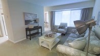 This single-floor residence offers a cohesive, open layout centered around a well-appointed living area. The living room is finished with soft wall-to-wall carpeting in a warm neutral tone and painted in a calming light blue-gray, creating a serene and inviting atmosphere. A large window fitted with horizontal blinds and accented by floor-length drapes allows abundant natural light to fill the space throughout the day.

The living area is thoughtfully arranged, featuring a central white coffee table paired with patterned upholstered seating, including a sofa and accent chair with coordinating decorative pillows. A dark-finished console table along one wall provides additional display and storage space, topped with a table lamp and decorative accessories. A landscape artwork piece above the console adds visual interest to the room.

An open sightline connects the living room to an adjacent dining area, visible in the background and finished in consistent neutral tones with natural light filtering through. The overall design palette throughout the space relies on soft blues, grays, and warm beiges, lending the home a calm, coastal-inspired aesthetic well-suited to comfortable, everyday living.