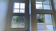 This two-storey space makes a striking architectural statement with its double-height ceiling, creating an expansive, open atmosphere that feels both grand and airy. The most defining feature of the main living area is its dramatic wall of windows, which spans two levels. Large lower windows are paired with upper clerestory windows, together allowing an exceptional amount of natural light to pour into the interior throughout the day. The neutral, warm-toned walls complement the clean lines of the window frames, contributing to a modern and understated aesthetic. Views through the windows reveal a verdant, tree-lined outdoor setting, providing a natural backdrop that enhances the sense of calm and connection to the outdoors. The generous vertical volume created by the double-height design adds a sense of grandeur rarely found in typical residential spaces. Overall, the architecture prioritizes light, openness, and visual connection to the surrounding landscape, making this a distinctly contemporary and thoughtfully designed space.