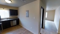 This single-story space features a practical, straightforward layout with light wood-style laminate flooring that runs throughout the visible areas, creating a cohesive and easy-to-maintain surface underfoot. The kitchen area is equipped with dark espresso-toned cabinetry that provides a strong contrast against the neutral, light-colored walls. A stainless steel dishwasher is integrated into the cabinetry, contributing to the functional nature of the kitchen. The walls are finished in a clean, off-white tone that keeps the space feeling bright and open. A flush-mounted electrical panel is centrally located on the main wall, offering convenient utility access. Ceiling-mounted HVAC vents are visible, indicating a central air system throughout. White baseboards frame the flooring transitions cleanly. A doorway with dark trim leads into an adjacent room, which features carpeted flooring, suggesting a bedroom or secondary living area. The overall design is utilitarian and move-in ready, with a neutral palette that provides flexibility for personalization.