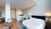 This thoughtfully designed studio integrates living, sleeping, and cooking functions within a single, open-plan layout. The sleeping area anchors one end of the space, centered around a generously sized bed with a tufted navy blue headboard, crisp white bedding, and accent pillows. A patterned area rug adds visual warmth underfoot, complementing the light wood-look flooring that runs throughout the entire unit.

The kitchen occupies a defined corner, featuring light yellow upper cabinetry, a clean white lower peninsula with bar seating, and stainless steel appliances including a refrigerator and microwave. The peninsula doubles as a casual dining counter, accompanied by a small round table and mixed seating — ideal for everyday meals.

Recessed lighting and a track lighting system along the ceiling provide ample illumination throughout the space, while the neutral wall tones in white and soft grey contribute to an airy, open atmosphere. A large closet near the entry adds practical storage. The overall design balances functionality with a modern, welcoming aesthetic suited to everyday living.