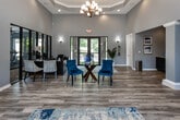 Located at 424 N Cedar Bluff Rd in Knoxville, Tennessee, this single-floor community clubhouse and leasing center offers an elegant and well-appointed environment designed to impress. The main reception area commands attention with soaring tray ceilings adorned by a statement chandelier, recessed lighting, and expansive light gray walls that lend an airy, sophisticated atmosphere. Wide-plank hardwood-style flooring runs throughout, complementing the blue and neutral accent furnishings that give the space a cohesive, modern feel. A central seating arrangement—featuring a glass-topped table surrounded by rich blue velvet chairs—serves as an inviting focal point for the open main area. Large black-framed glass doors at the entrance flood the interior with natural light and offer views of lush greenery outside. Decorative framed artwork and accent lighting punctuate the walls, adding warmth and character. A dedicated office workspace, separated by floor-to-ceiling glass partitions, provides a professional leasing area with direct visual access to a fully equipped fitness center beyond, which features exercise machines set against a bold blue accent wall. Decorative columns and an adjacent kitchenette area with dark cabinetry complete the interior, offering both function and style in a cohesive, well-designed package.