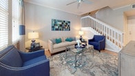 This well-designed two-story home offers a cohesive coastal-inspired aesthetic throughout, with light hardwood flooring, neutral gray walls, and crisp white trim creating a bright and welcoming atmosphere. The main level features an open-concept layout where the kitchen, dining area, and living room flow seamlessly together. The kitchen is outfitted with white shaker cabinetry, quartz countertops, stainless steel appliances, a gray subway tile backsplash, and a central island with bar seating — ideal for both meal preparation and casual dining. Adjacent to the kitchen, the dining area accommodates a full glass-top table and benefits from natural light through plantation-shuttered windows framed by navy and cream striped curtains. The living room impresses with vaulted ceilings, a ceiling fan, crown molding, and a white-railed staircase leading to the upper floor. Upstairs, the bedrooms provide comfortable retreats. One bedroom features sliding glass doors opening to a private balcony with outdoor views, while another includes a walk-in closet. A full bathroom on the upper level offers a soaking tub, tile flooring, and an extended vanity with white cabinetry and quartz countertops. The space combines modern finishes with a relaxed, coastal character throughout.