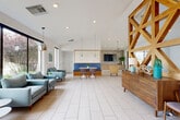 Located at 4221 E Ray Rd in Phoenix, AZ, this single-floor space showcases a thoughtfully designed mid-century modern aesthetic across an open, flowing layout. Light-toned large-format tile flooring runs throughout, complementing crisp white walls and an abundance of natural light. A striking geometric wood lattice partition serves as a dramatic architectural focal point, visually dividing the living and dining areas while maintaining a sense of openness. The living area features floor-to-ceiling windows that frame outdoor views of lush desert landscaping, with pendant lighting adding a warm, contemporary touch. The dining area centers around a live-edge wood table paired with upholstered mid-century chairs, beneath a cluster of globe pendant lights. A full glass curtain wall floods the space with natural light and connects visually to an adjacent workspace. The kitchen is appointed with warm wood cabinetry, a white quartz island with bar seating, a gas cooktop, stainless steel refrigerator, and a distinctive seafoam-green subway tile backsplash. A pass-through window connects the kitchen to the main space, enhancing its communal feel. French doors provide additional access points, and the overall ambiance is bright, airy, and inviting throughout.