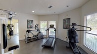This single-story fitness facility is thoughtfully designed to accommodate a comprehensive workout experience. The space is divided into distinct functional zones, each tailored to a specific type of exercise. The cardio area features multiple machines, including a treadmill, elliptical trainer, and additional cardiovascular equipment, all positioned near windows fitted with horizontal blinds that allow natural light to filter in while providing views of surrounding greenery. Motivational fitness posters accent the white walls, adding visual interest to the space.

The strength training area is anchored by a substantial multi-station cable weight machine finished in white, offering a variety of resistance exercises. Floor-to-ceiling mirrors line one wall, visually expanding the space and allowing users to monitor their form during workouts. The dark, durable flooring throughout provides a practical and resilient surface suited to heavy equipment and high-traffic use.

A French door with glass panes connects to an adjacent room, visible through a pass-through window, suggesting an office or reception area. Recessed lighting and textured ceilings complete the interior. The overall layout is open and functional, providing ample room to move between equipment comfortably.